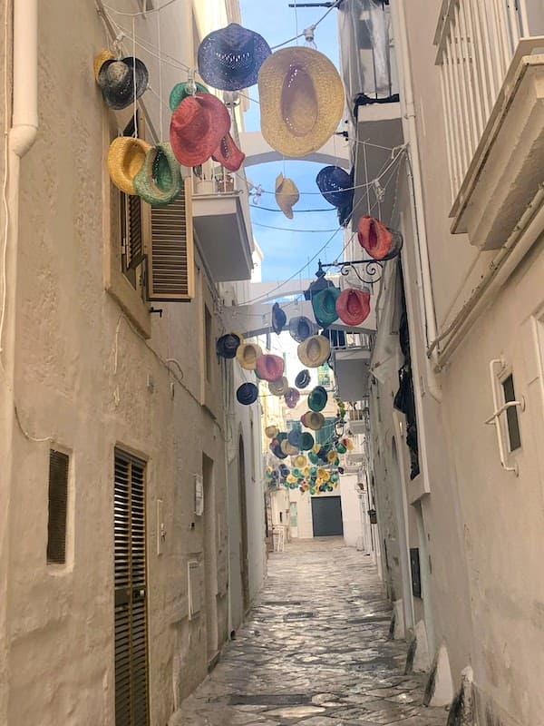 I Drove All Over Puglia, Italy — Here Are 6 Road Trip Itineraries That Actually Work Stone-paved alley with colourful hanging straw hats in the historic old town of Monopoli, Puglia, Italy.