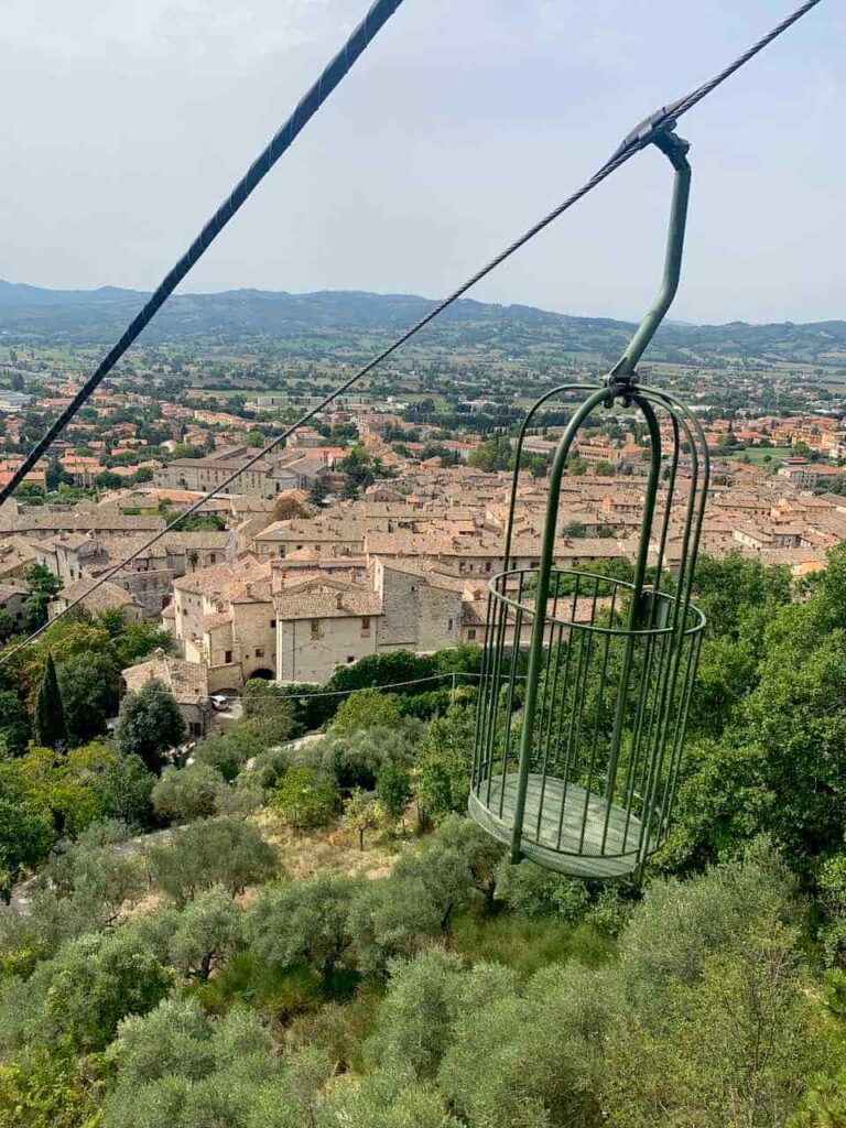 Green open-air birdcage cabin of the Funivia Colle Eletto cable car rising above the medieval town of Gubbio, Italy
